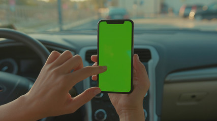 Lviv, Ukraine - May 19, 2018: Close-up of young female driver showing vertical smartphone doing tapping gestures on mock-up green screen inside modern automobile.