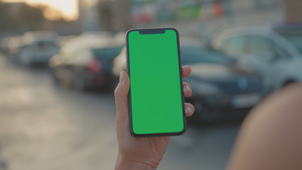 Lviv, Ukraine - May 19, 2018: Close-up of young urban girl showing vertical smartphone with greenscreen chrome key technology on parking lot outside. City life and modern gadgets.