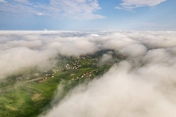 Aerial view of white clouds above a town or village with rows of buildings and curvy streets between green fields in summer. Countryside landscape from above.