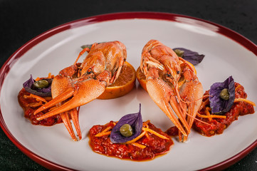 Close up boiled crayfish. Delicacy with chili pepper sauce, orange, zest, basil and capers on a plate. Gourmet cuisine. Dark concrete background