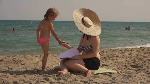 Serious woman in a swimsuit and straw hat shows a girl how to draw watercolor sketches. Lesson of drawing and sketching on the seashore. Children learn to paint in watercolor