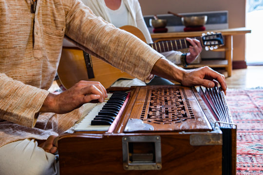 Closeup of male shaman hands playing sacred kirtan music with fingers on keys of harmonium with woman playing classical guitar in room to meditate