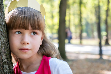 Portrait of a pretty child girl in a paper crown on her head leaning to a tree in summer park.