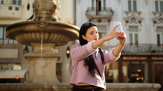 Young woman is sitting on fountain in historical part of Bratislava city and taking selfie with smartphone in SLOW MOTION HD VIDEO. Low depth of field and blurred background. Half speed.