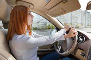 Young redhead woman driver behind a wheel driving a car smiling happily.