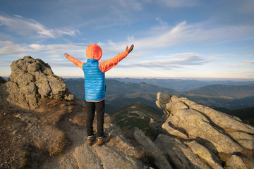Young child boy hiker standing with raised hands in mountains enjoying view of amazing mountain landscape at sunset.