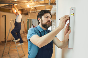 Portrait of bearded construction worker measuring level of wall while renovating house, copy space