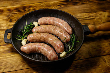 Raw sausages ready for preparation with rosemary, garlic and spices in cast iron grill frying pan on wooden table