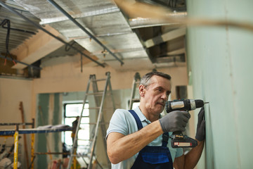 Side view portrait of senior construction worker drilling wall while renovating house alone, copy space
