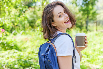Happy positive student girl with backpack smiling on green park background. Woman with take away coffee cup having rest in campus during lunch break. Education and leisure concept.