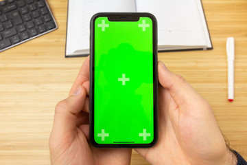Mockup image of hand holding black mobile phone with green screen in the office table on the background