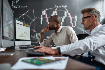 Planning for secure future. Two diverse colleagues traders discussing ideas while sitting in the office in front of multiple computer screens. Blackboard full of data analyses in the background.
