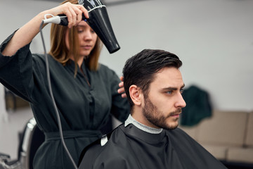 Handsome blue eyed man sitting in barber shop. Hairstylist Hairdresser Woman cutting his hair. Female barber.