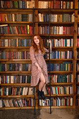 young beautiful red-haired girl in a dress stands on the background of the library and books in the apartment in the loft style