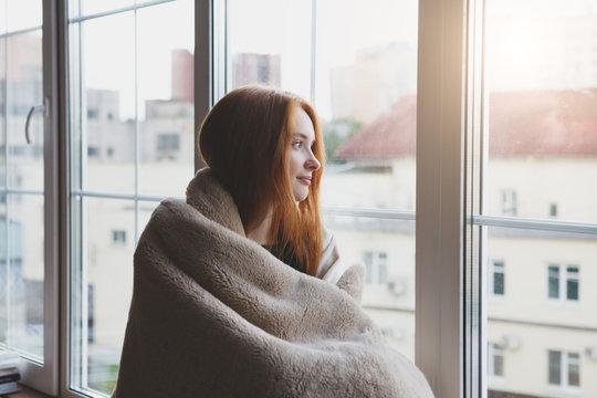 beautiful thoughtful pensive calm smiling young woman looking through the window at home wrapped in warm comfy blanket