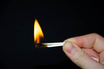 A hand holds a burning match on a black background. A wooden match burns in the hands of a macro. Igniting a match on a box. Smoke from a match.  Flash