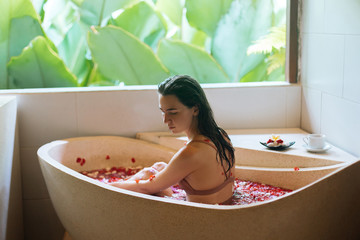 Back view of Woman relaxing in stone bath tube with tropical flowers with jungle view, organic skin care, luxury spa hotel, Bali Indonesia