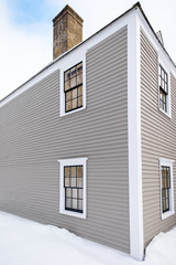 The corner view of a two storey vintage house. The exterior is grey colored clapboard with white trim. There are multiple windows made of glass and black edging. There's a brick chimney in building.