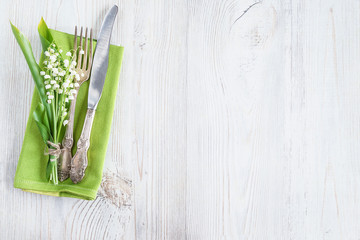 Festive table setting. Vintage fork and knife, bouquet of lilies of the valley, green napkin on white wooden background. Space for text, flat lay