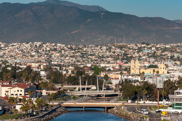 Fototapeta premium Ensenada, Mexico - January 17, 2012: Arroyo Ensenada empties in bay and leads to Yellow Cathedral of Our Lady of Guadalupe under evening sun emersed in cityscape and black moutain as backdrop.
