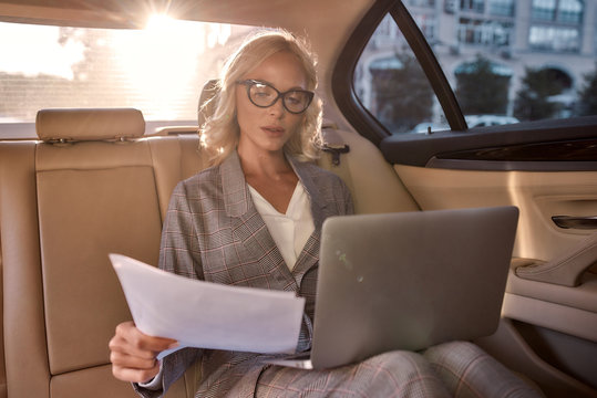 Self-made woman. Beautiful and successful business woman in full suit analyzing documents and working on laptop while sitting in the car