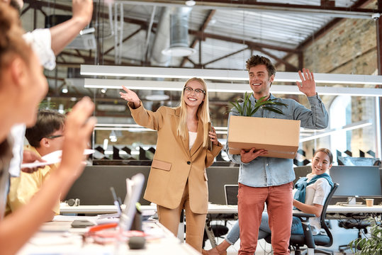 Happy to be in your team. Young handsome man in casual wear holding box with things and waving to his new coworkers while standing in the modern office