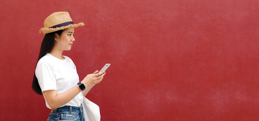 Happy asian girl using mobile phone standing over red wall background with copy space, Young asia...
