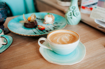 A Cup of coffee in the color of blue gradient from white to blue full of coffee with milk foam and a pattern on the background of dessert chocolate fondan at the table of a cozy light cafe. Lifestyle