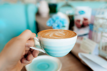 a light-skinned hand holds a delicate porcelain mug of gradient blue full of coffee with a milky froth and pattern. Breakfast in the city cafe.