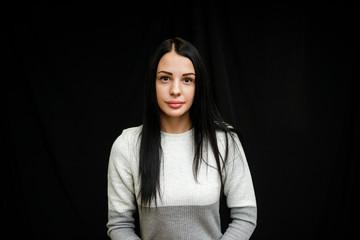 Portrait of serious beautiful female with black hair, has minimal makeup, looks calmly at camera, wears white jumper, stands against black background, being deep in thoughts