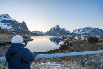 Woman with winter clothes standing on the bridge overlooking the fishing village of Reine in Lofoten during cold weather and blue hour. Red rorbuer infront and snow covered peaks in the back.