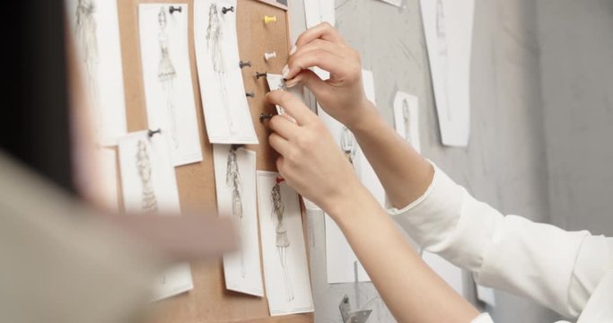 Asian female clothes designer is looking at her sketches pinned to wall in office, looking for inspiration for her new collection - fashion concept 4k footage