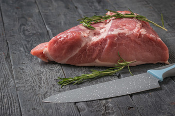 A large piece of pork with rosemary and a knife on a black wooden table.