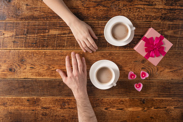 Valentine's Day celebration concept. A nice gift for your loved one. Hands of man and woman with coffee mugs on a wooden table background. Copy space. Flat lay. Close-up.