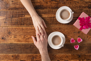 Valentine's Day celebration concept. A nice gift for your loved one. Hands of man and woman with coffee mugs on a wooden table background. Copy space. Flat lay. Close-up.