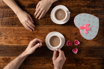 Valentine's Day celebration concept. A nice gift for your loved one. Hands of man and woman with coffee mugs on a wooden table background. Copy space. Flat lay. Close-up.