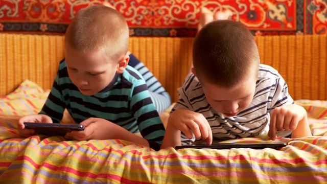 funny boys are lying on couch to play network computer games on smartphone and tablet. Parents gave their children new gadgets. Modern children are well aware of latest technology. Brothers are happy