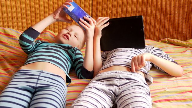 funny boys are lying on couch to play network computer games on smartphone and tablet. Parents gave their children new gadgets. Modern children are well aware of latest technology. Brothers are happy