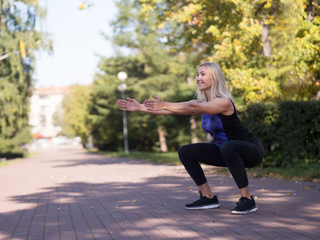 Caucasian smiling middle-aged athletic woman doing deep squat on footway in urban park, outdoor,...