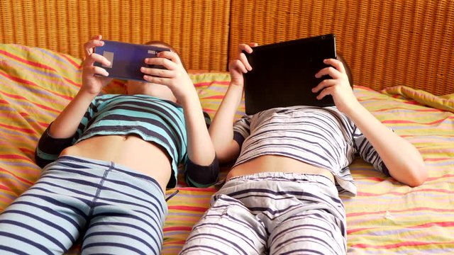 funny boys are lying on couch to play network computer games on smartphone and tablet. Parents gave their children new gadgets. Modern children are well aware of latest technology. Brothers are happy