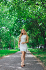 Naklejka na ściany i meble Portrait of a charming blond woman wearing beautiful white dress standing on the road under trees.