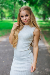 Naklejka premium Portrait of a charming blond woman wearing beautiful white dress standing on the road under trees.