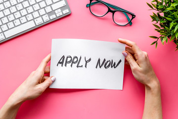 Apply now. Advertisement in hands on pink office desk top-down