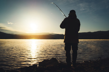 silhouette fisherman with fishing rod at sunrise sunlight, outline man enjoy hobby sport on evening lake, person catch fish at night sky, relaxation fishery concept