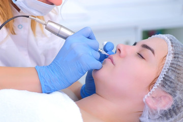 Naklejka premium Cosmetologist is applying permanent makeup on woman lips using tattoo machine in clinic. Beautician and patient face closeup. Lips microblading procedure for girl. Beauty industry concept.