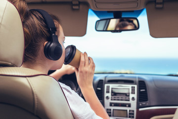 Woman driver in the headphones driving a car. Girl relaxing in auto trip drinking coffee paper cup traveling along ocean tropical beach in background. Traveler concept.
