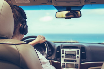 Fototapeta na wymiar Woman driver in the headphones driving a car. Girl relaxing in auto trip traveling along ocean tropical beach in background. Traveler concept. Back view