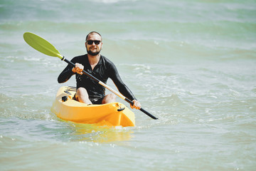 Handsome smiling young man in sunglasses enjoying kayaking in warm sea on weekend