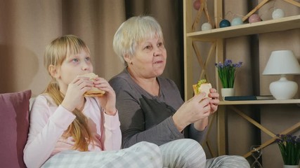 Active elderly woman with granddaughter together watching TV and eating sandwiches