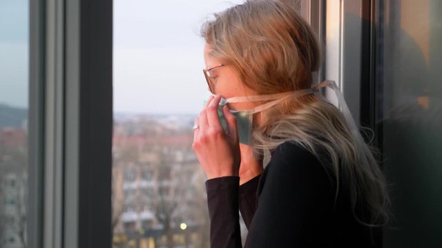 young woman with medical mast stand in front of the window and watching sunset and cough as ill